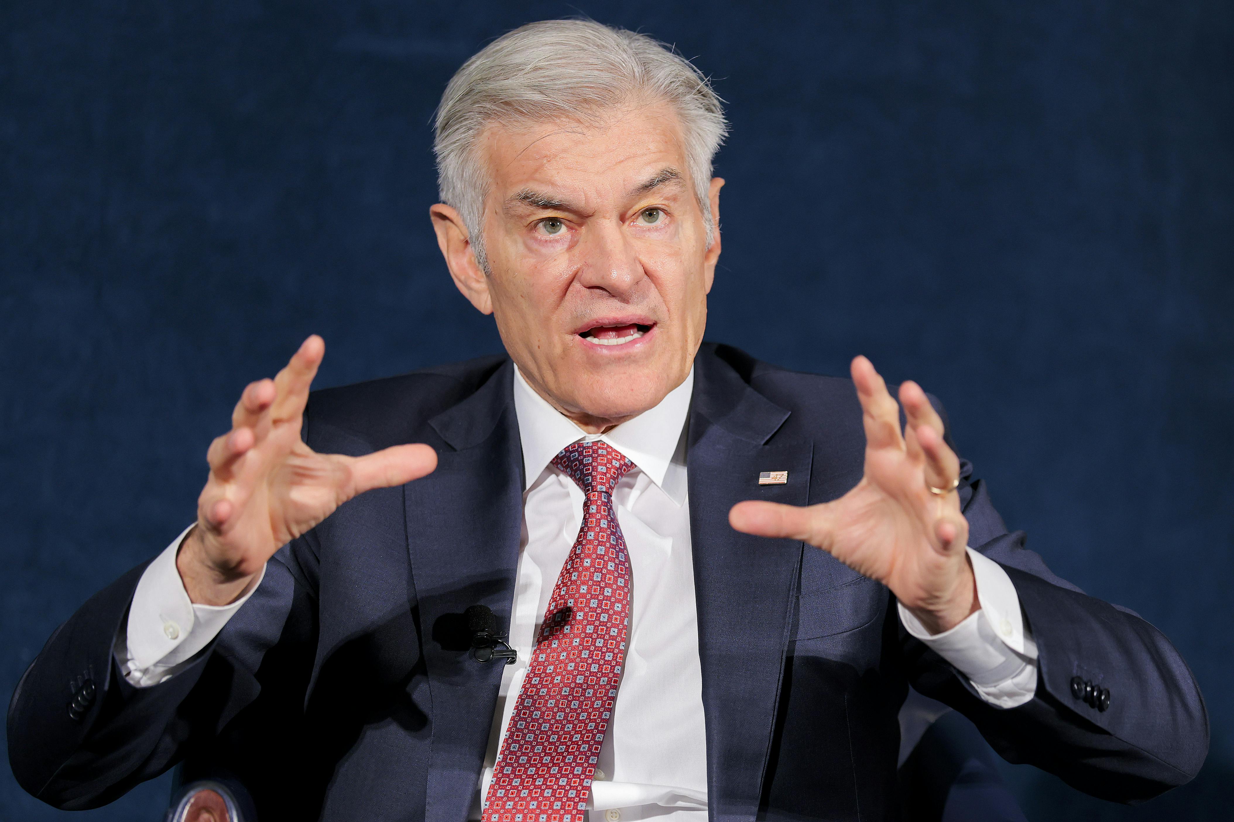 Dr. Oz is seen from the waist up, speaking while seated and raising both hands while talking.