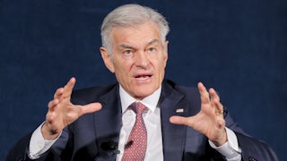 Dr. Oz is seen from the waist up, speaking while seated and raising both hands while talking.