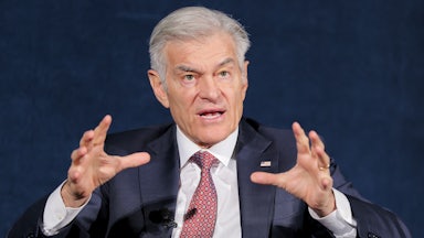 Dr. Oz is seen from the waist up, speaking while seated and raising both hands while talking.