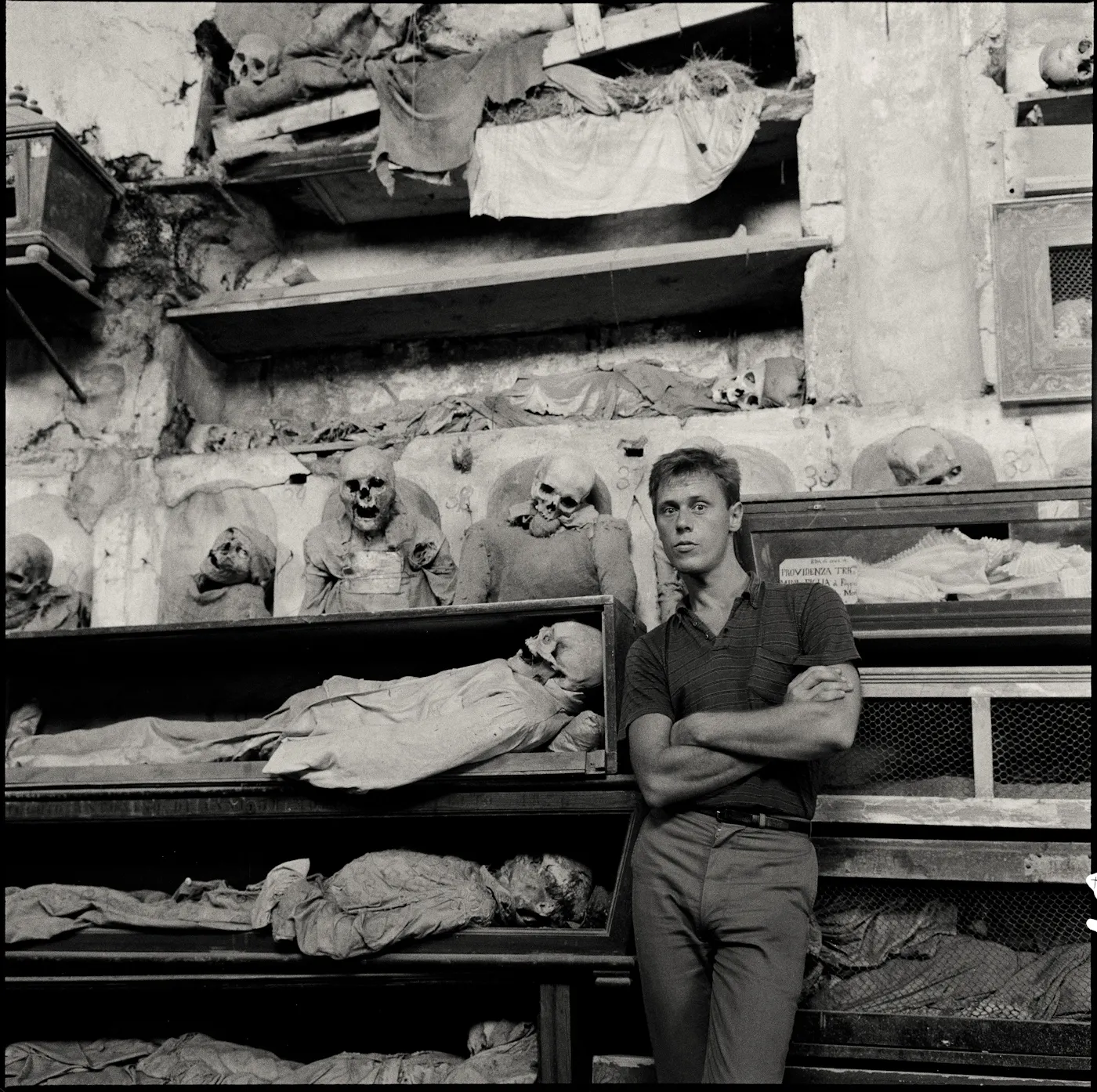 A 1963 black and white photograph by Peter Hujar of Paul Thek i at the Capuchin Catacombs n Sicily where he is standing in front of mummified bodies.