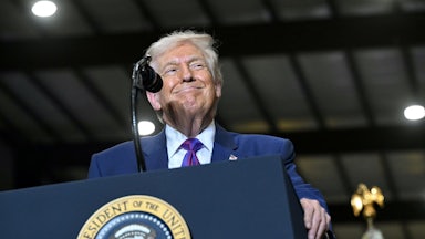 Donald Trump smiles while wearing a blue suit leans over a podium at an angle with the presidential seal partially visible.