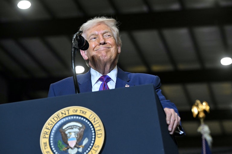 Donald Trump smiles while wearing a blue suit leans over a podium at an angle with the presidential seal partially visible.