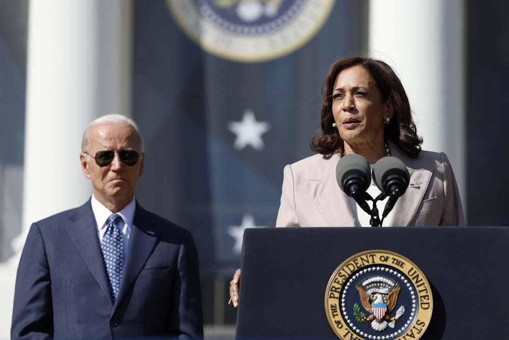 Kamala Harris speaks at a podium with Joe Biden, wearing sunglasses, behind her.