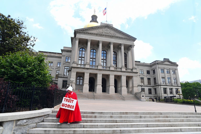 A woman wearing an outfit from The Handmaid's tale stands on the steps of the Georgia Capitol, holding a sign that reads "Trust Women."