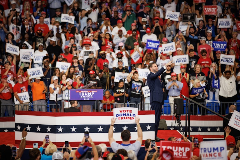 People at a Donald Trump rally hold up signs that say “You’re Fired!” as J.D. Vance stands onstage and points at the crowd