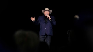 Jeff Rath speaks into a microphone on a dark stage while wearing a cowboy hat.