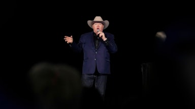 Jeff Rath speaks into a microphone on a dark stage while wearing a cowboy hat.