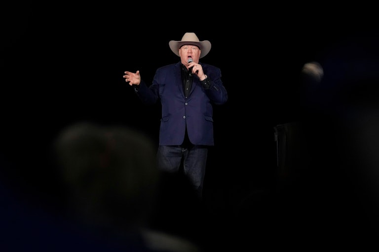 Jeff Rath speaks into a microphone on a dark stage while wearing a cowboy hat.