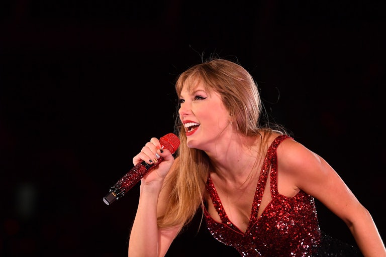 Taylor Swift laughs as she performs on stage, bending forward as she holds a mic in her hand