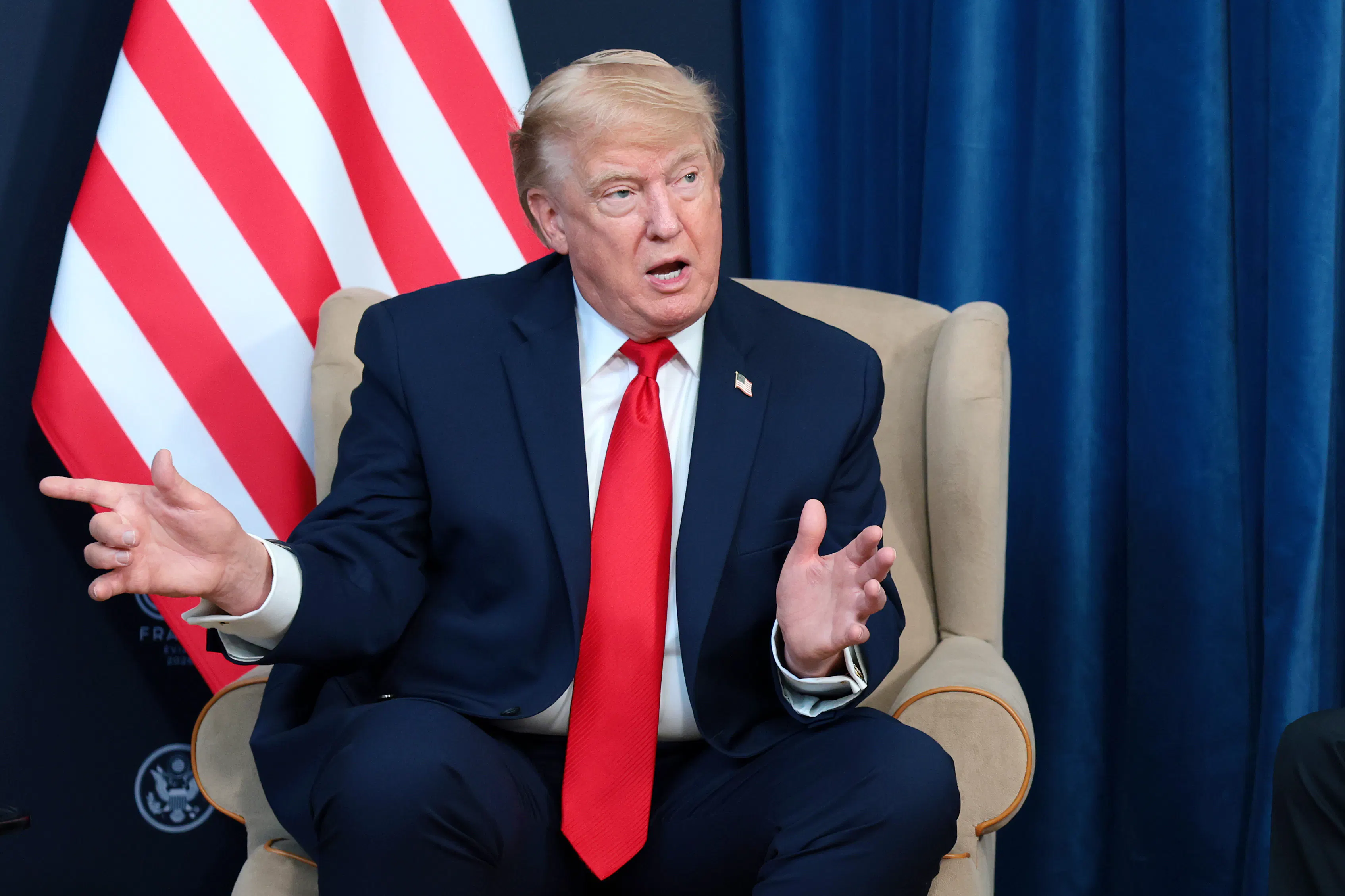 Donald Trump speaking while sitting in front of a U.S. flag