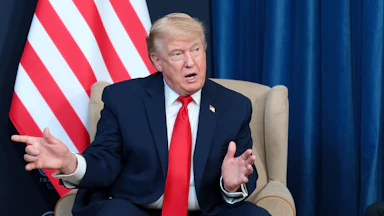Donald Trump speaking while sitting in front of a U.S. flag