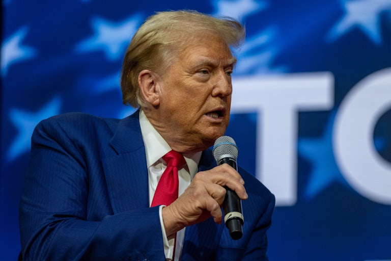 Donald Trump, seated, holds a mic and slighty leans over while talking to someone not shown on camera