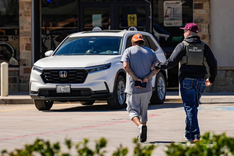 A DPS special agent steers a handcuffed brown man with tattoos on his arms.