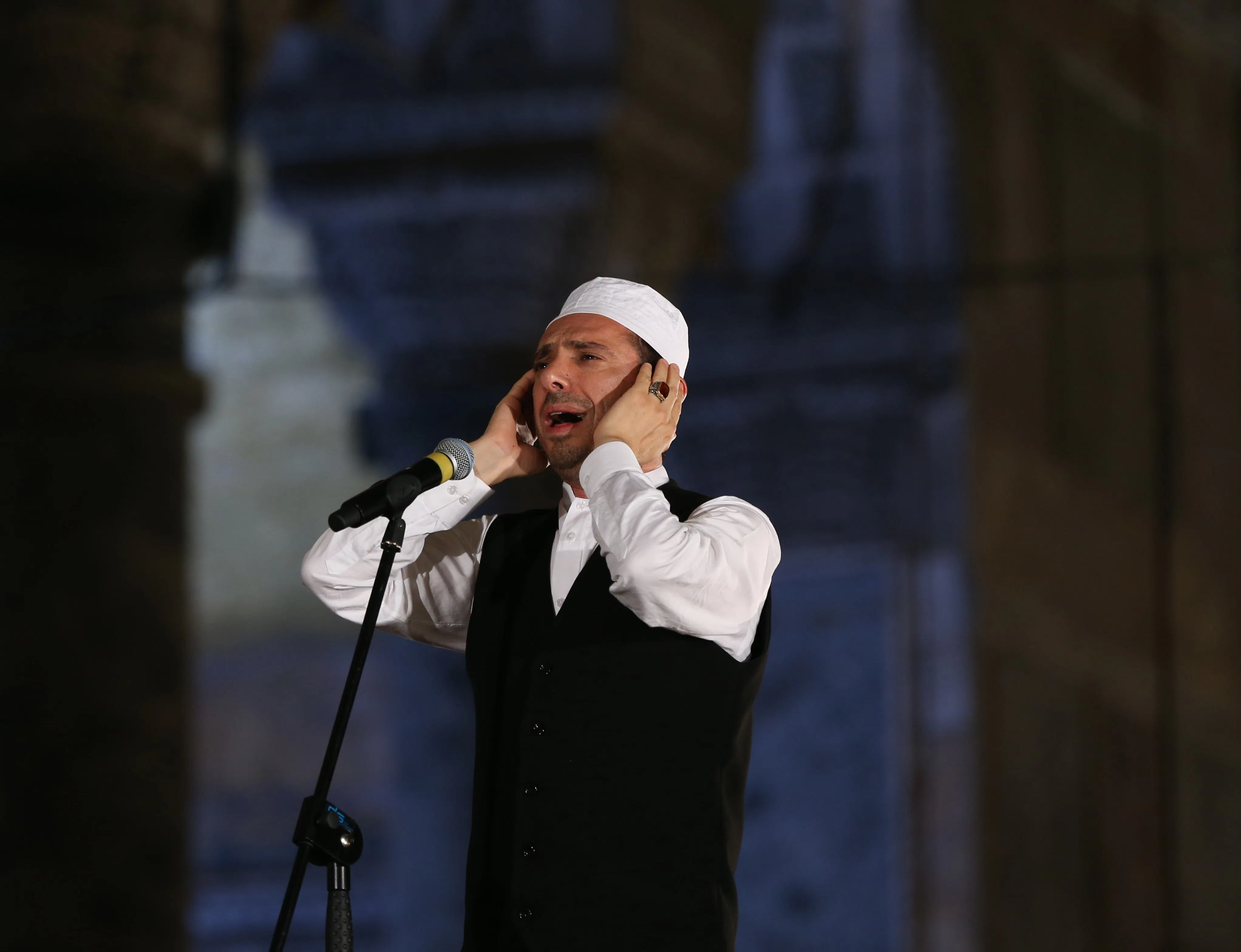 A man wearing a black vest, white dress shirt, and white Islamic cap stands in front of a microphone, his mouth open with his hands at his ears. 