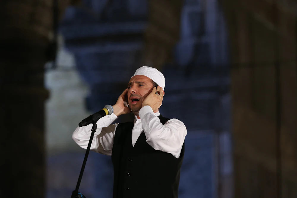 A man wearing a black vest, white dress shirt, and white Islamic cap stands in front of a microphone, his mouth open with his hands at his ears.