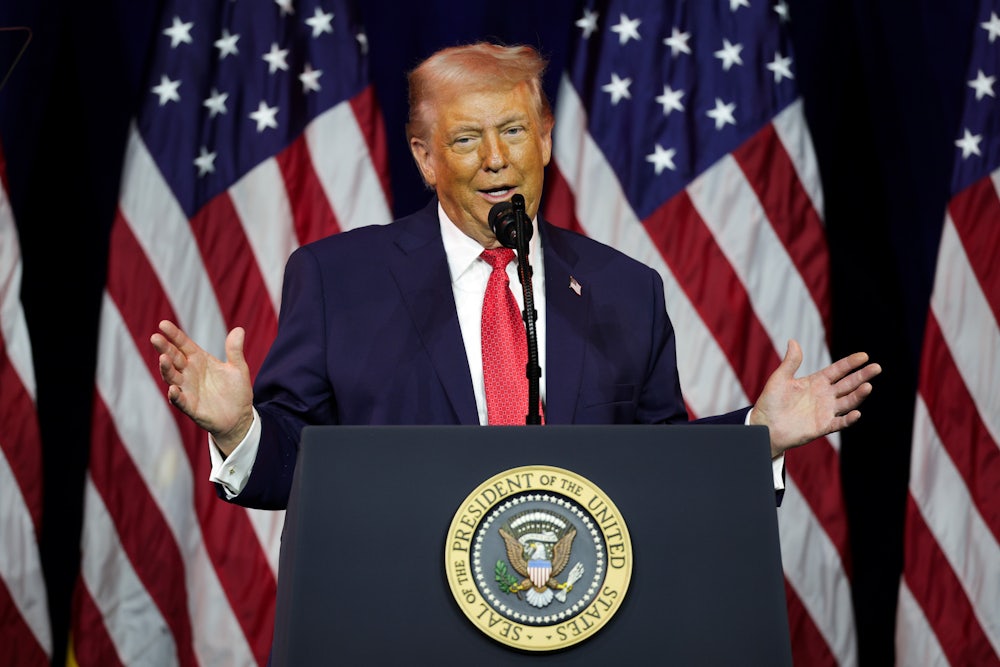 Donald Trump holds his hands out while speaking at a podium, in front of an array of American flags.
