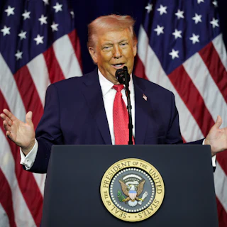 Donald Trump holds his hands out while speaking at a podium, in front of an array of American flags.