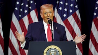 Donald Trump holds his hands out while speaking at a podium, in front of an array of American flags.