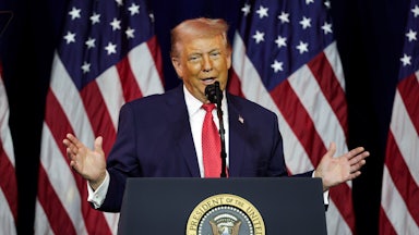 Donald Trump holds his hands out while speaking at a podium, in front of an array of American flags.