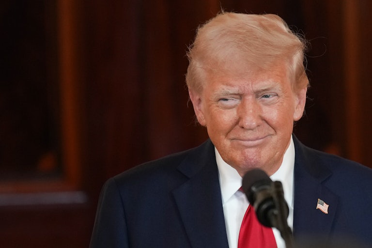 Donald Trump smiles with his mouth closed while standing before a mic.