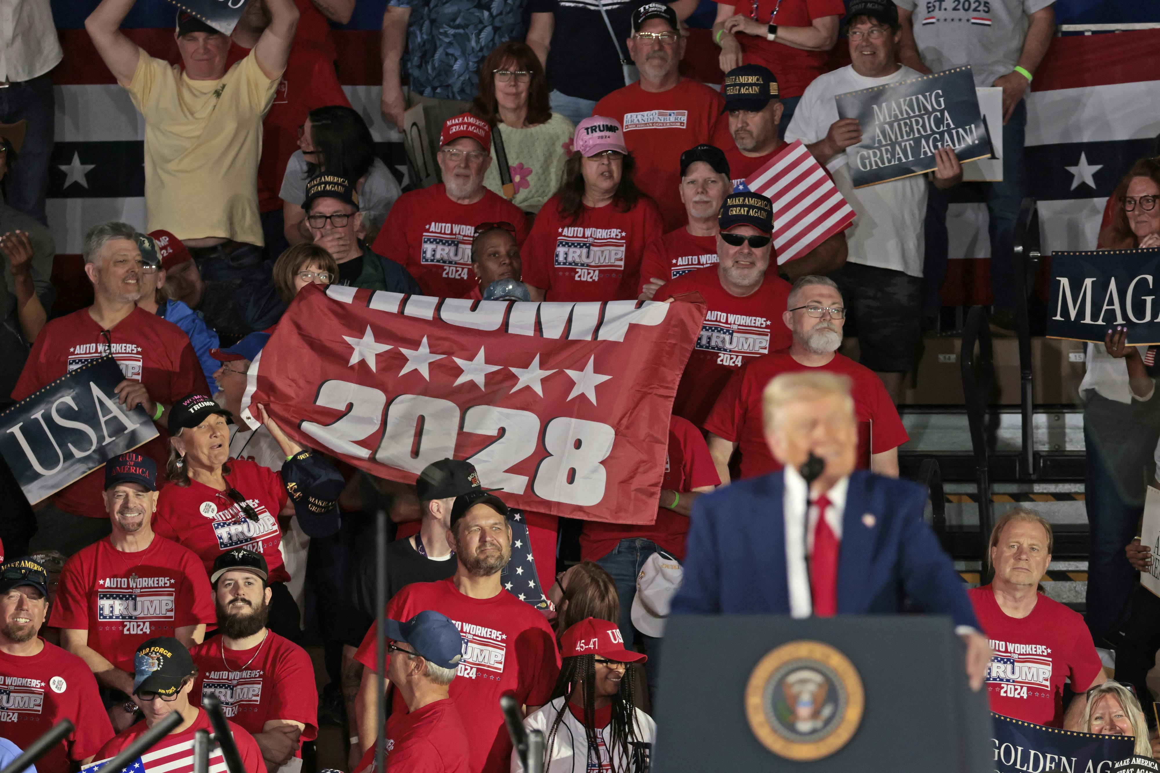 Several people in a crowd hold a flag that reads "Trump 2028" as Trump speaks.