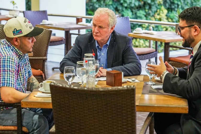 Senator Chris Van Hollen sits at the table with wrongly deported immigrant Kilmar Abrego Garcia. On the table are two glasses filled with a clear liquid and cherries on the rim. Another man sits at the table with them.