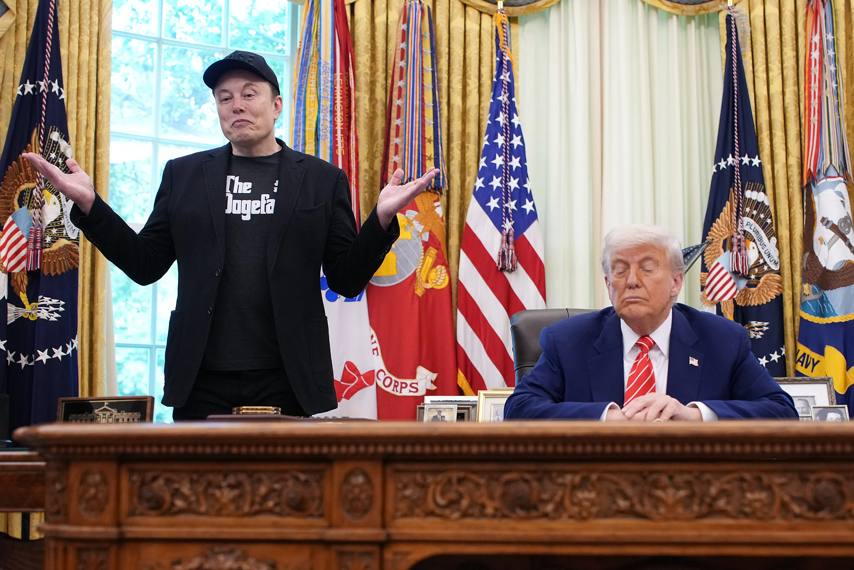 Elon Musk shrugs while standing next to the Resolute Desk. Donald Trump sits at the desk, looking down.