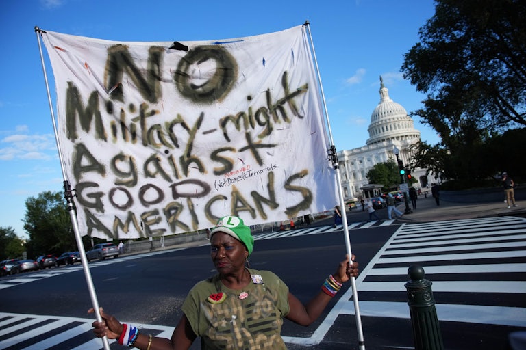 A Black woman wearing a Luigi cap and an embroidered pin stands near the Capitol holding a large sign reading "No Military-might Against Good Americans."