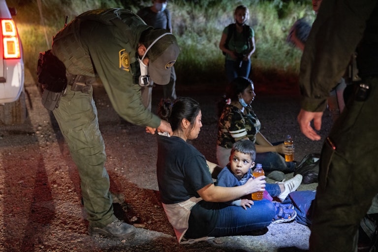 A Border Patrol agent bends down to place something on the neck of an undocumented immigrant sitting on the ground with a child in her lap.