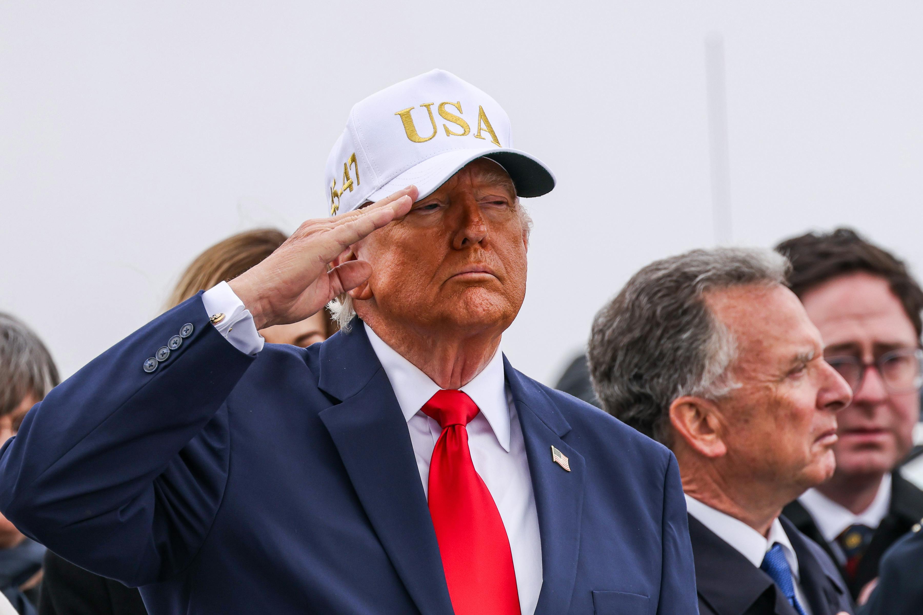 Donald Trump salutes while wearing a white baseball cap that says "USA"