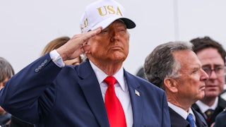 Donald Trump salutes while wearing a white baseball cap that says "USA"
