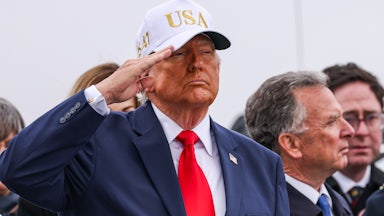 Donald Trump salutes while wearing a white baseball cap that says "USA"