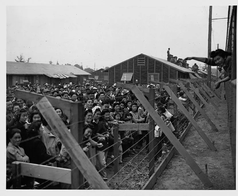 A Japanese American men, women, and children behind a barbed wire fence at a WWII era internment camp