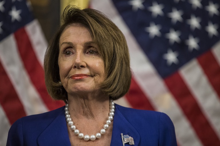 Nancy Pelosi, wearing a blue pantsuit and pearls, smiles and looks off into the distance in front of an American flag.