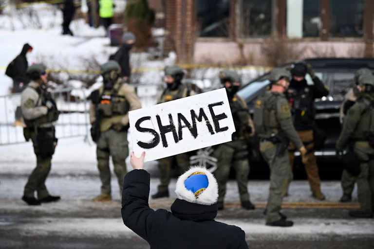 woman holds up sign reading "shame" in front of four ICE agents on