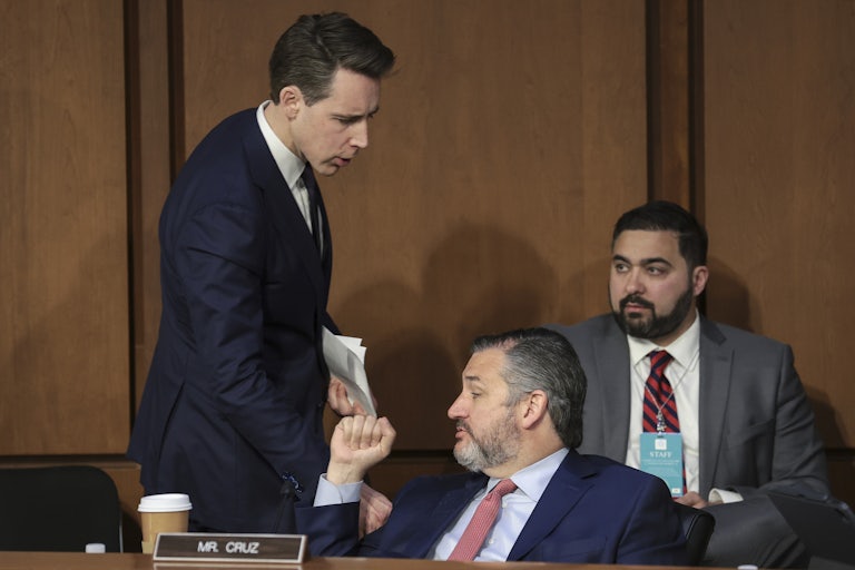 Josh Hawley bends down to talk to Ted Cruz, seated, who holds up a clenched fist