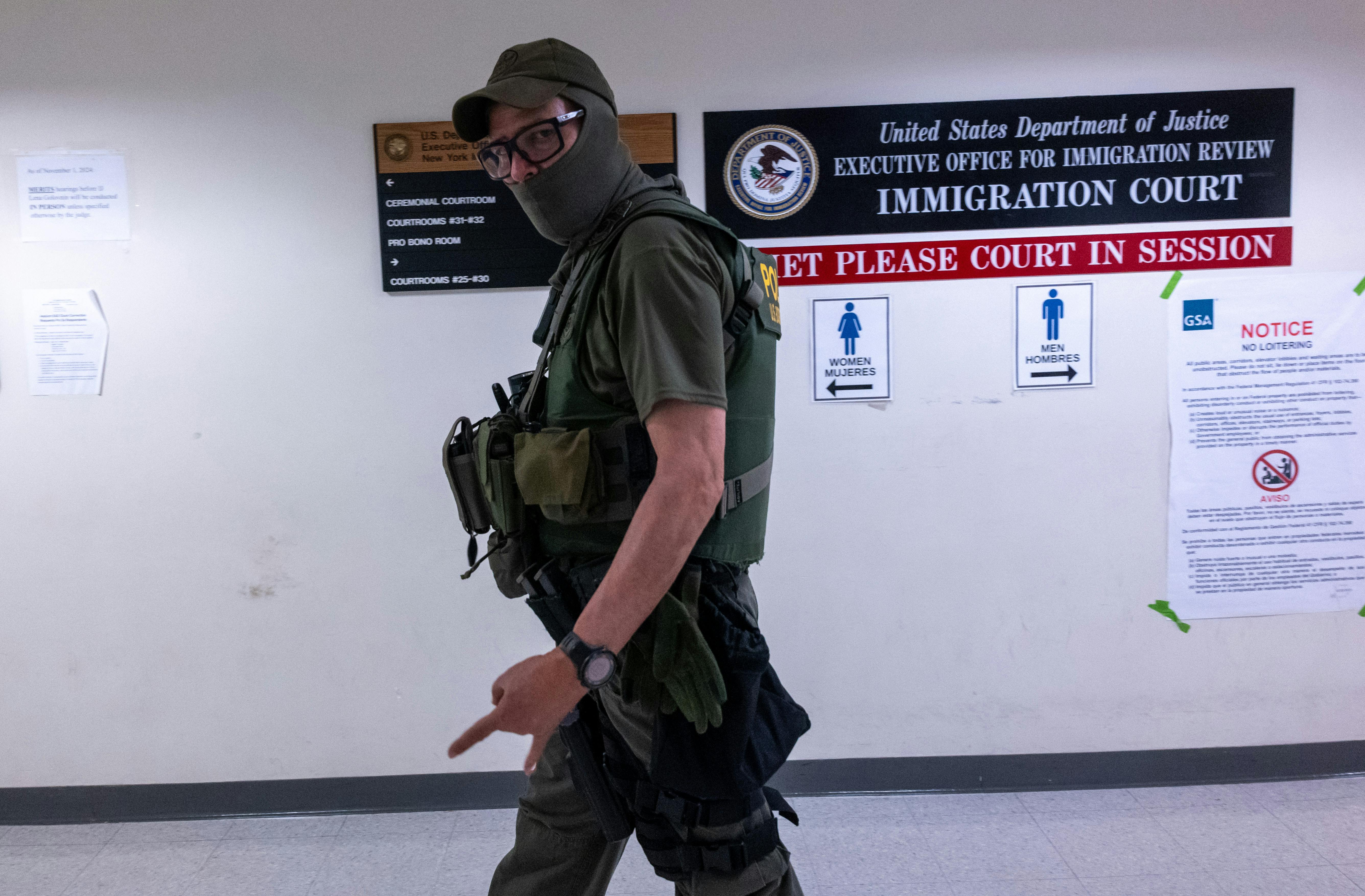 A white man wearing glasses, a face mask, and an ICE vest walks as he looks directly at the camera.