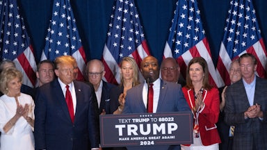 Tim Scott speaks at a podium, with Donald Trump beside him and others in the background.