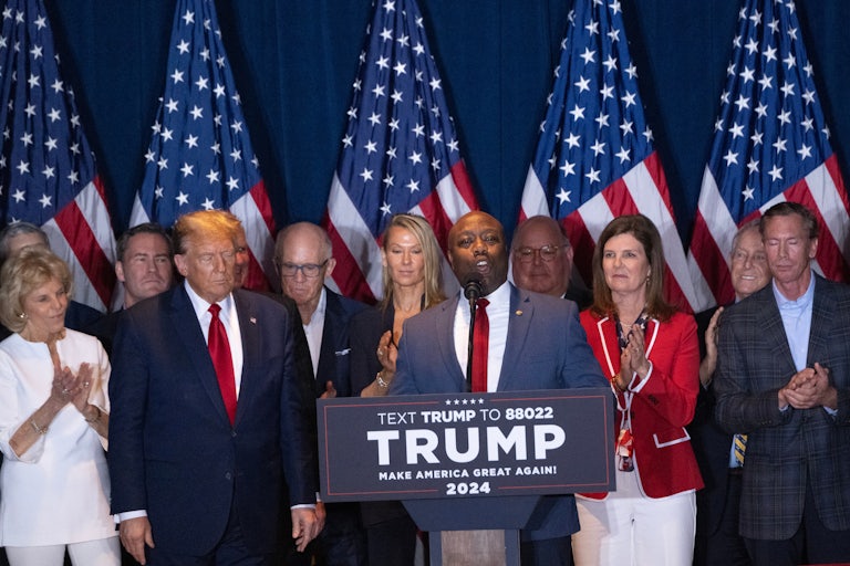 Tim Scott speaks at a podium, with Donald Trump beside him and others in the background.