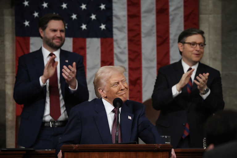 Donald Trump smiles weirdly while giving his speech to Congress. JD Vance and Mike Johnson stand and applaud in the background.
