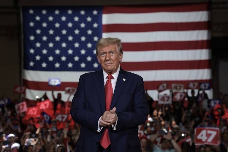 Donald Trump clasps his hands and smiles in front of a large US flag