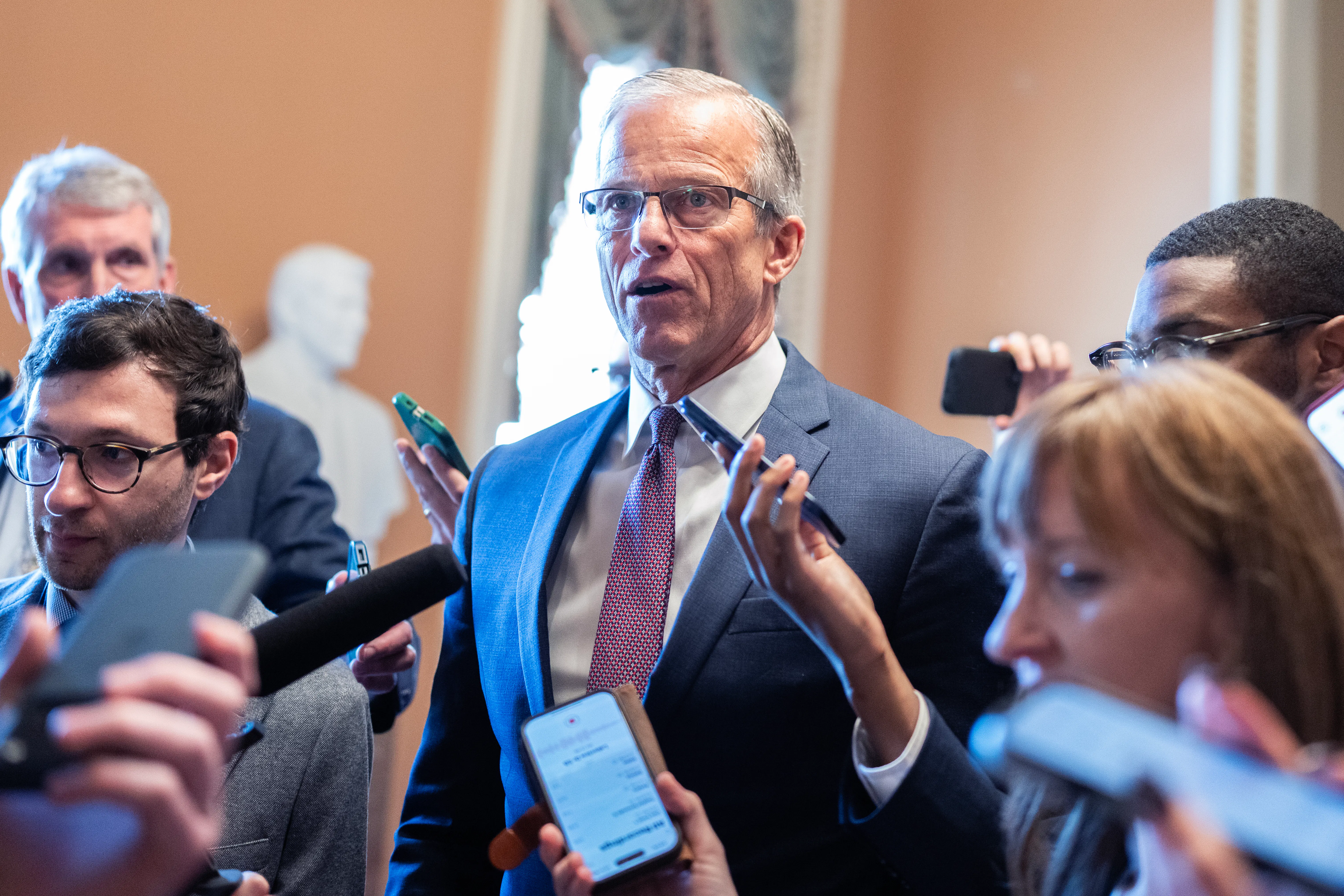 John Thune stands in a suit surrounded by people, some holding phones to record his words.