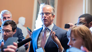 John Thune stands in a suit surrounded by people, some holding phones to record his words.