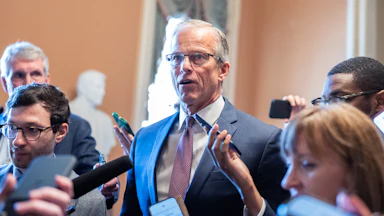 John Thune stands in a suit surrounded by people, some holding phones to record his words.