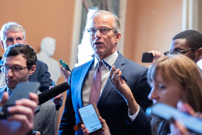 John Thune stands in a suit surrounded by people, some holding phones to record his words.