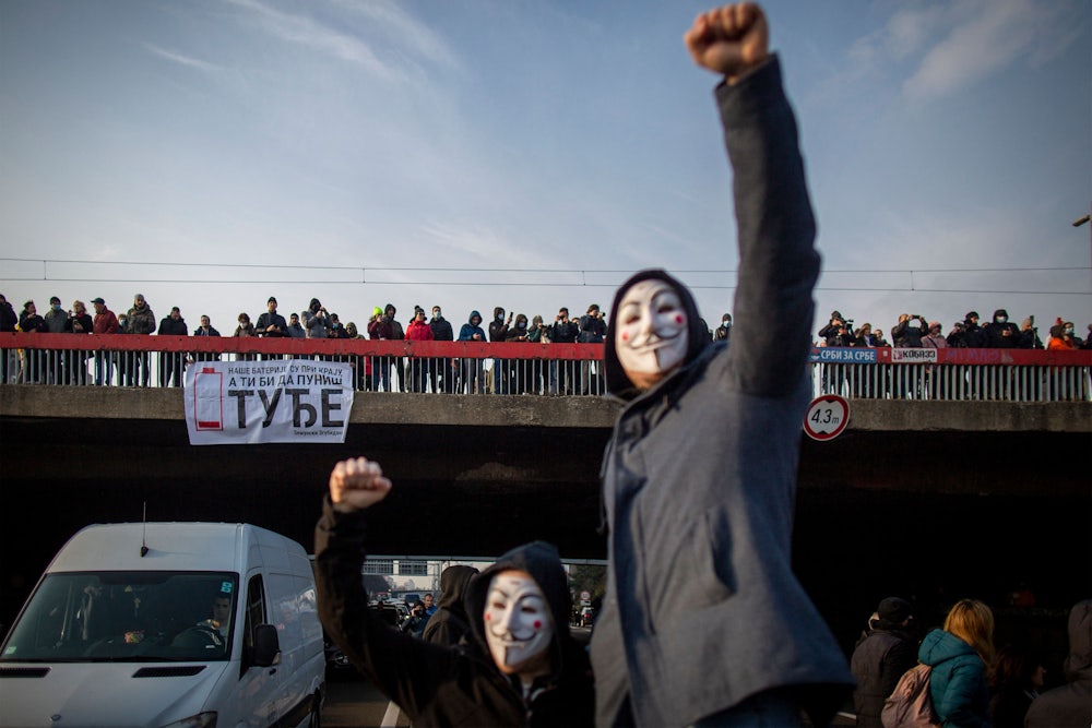 Two people with masks raise their fists, in front of an overpass crowded with people.