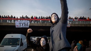 Two people with masks raise their fists, in front of an overpass crowded with people.