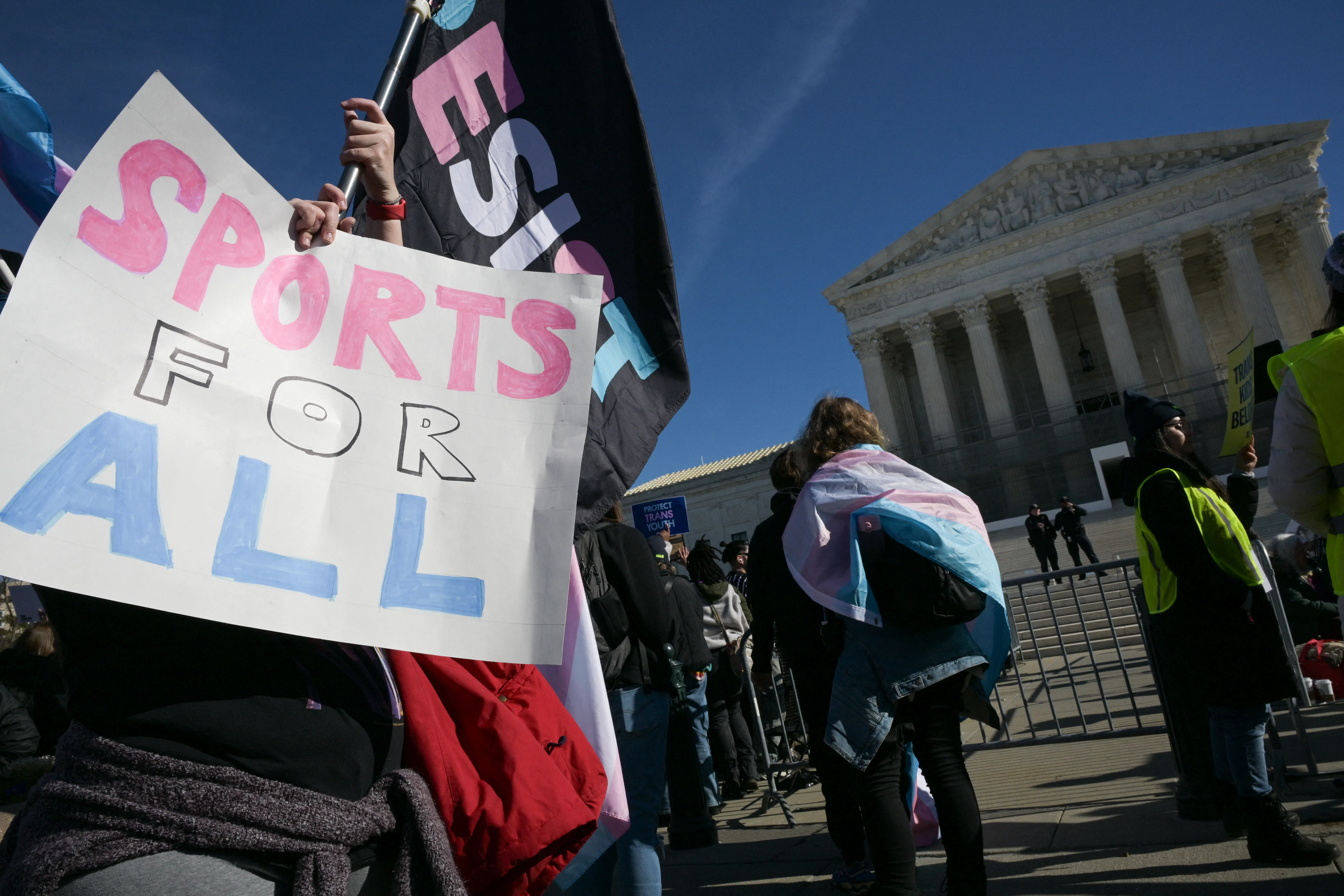 Someone holds up a sign that reads "Sports For All" while another waves a "Resist" flag. Both are in the trans flag colors and they stand before the Supreme Court.