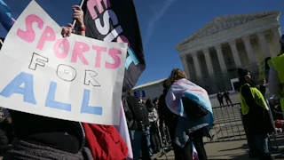 Someone holds up a sign that reads "Sports For All" while another waves a "Resist" flag. Both are in the trans flag colors and they stand before the Supreme Court.