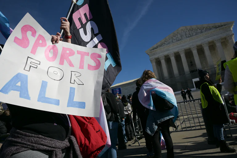 Someone holds up a sign that reads "Sports For All" while another waves a "Resist" flag. Both are in the trans flag colors and they stand before the Supreme Court.